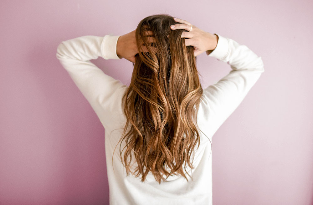 una mujer de espalda con su cabello rubio suelto y las manos puestas sobre su cabeza